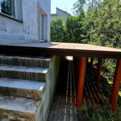 Nowy, wysoki taras na słupach, z drewnianą podłogą, sąsiaduje z betonowymi schodami i zielonym ogrodem.
