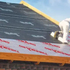 Pracownik montuje izolację na dachu, pokazując, jak ocieplić dach od zewnątrz. Materiał z napisem THERMANO "KEEP WARM" zapewnia komfort termiczny.
