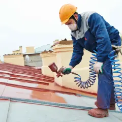 Pracownik w kasku maluje dach na czerwono, pomagając zdecydować, jaką wybrać farbę na dach.