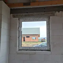 Ściana z bloczków z widocznym oknem i cofniętym nadprożem do rolet. W tle dom w budowie.