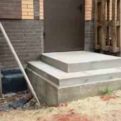Solidny fundament pod schody z betonu, gotowy na dalsze prace budowlane. Obok widać materiały budowlane i fragment domu.