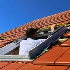 Monter montuje okno dachowe w blachodachówce, używając poziomicy. Praca na dachu pod błękitnym niebem.