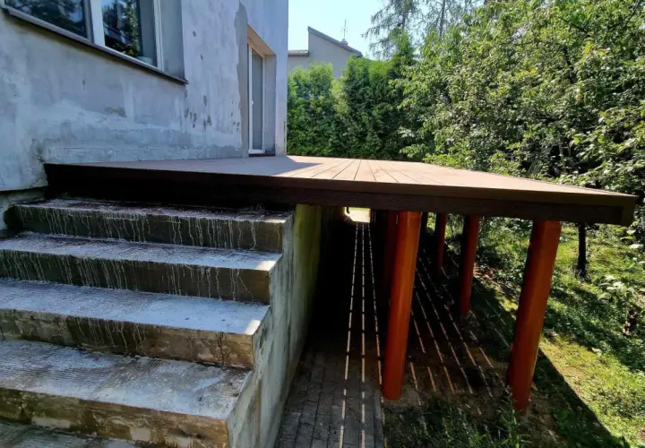 Nowy, wysoki taras na słupach, z drewnianą podłogą, sąsiaduje z betonowymi schodami i zielonym ogrodem.