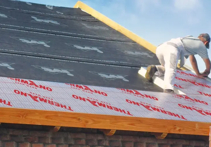 Pracownik montuje izolację na dachu, pokazując, jak ocieplić dach od zewnątrz. Materiał z napisem THERMANO "KEEP WARM" zapewnia komfort termiczny.