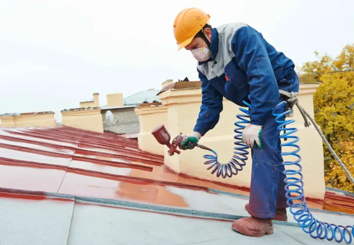 Pracownik w kasku maluje dach na czerwono, pomagając zdecydować, jaką wybrać farbę na dach.