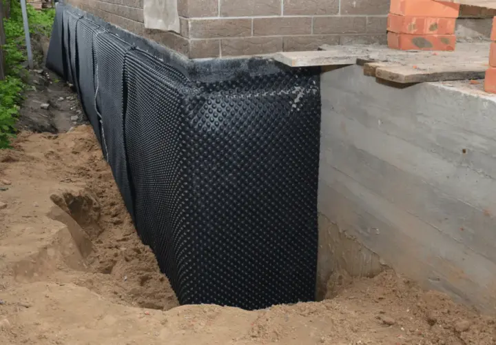 Folia kubełkowa chroni betonowy cokół przed wilgocią. Widać wykop i materiały budowlane.