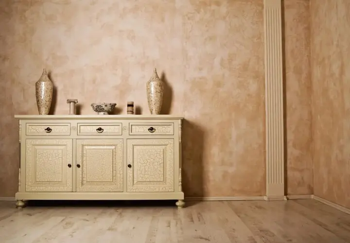 Elegancka komoda z crackle na drzwiach, inspirowana glinką wenecką, na tle ściany o ciepłej, rustykalnej fakturze.