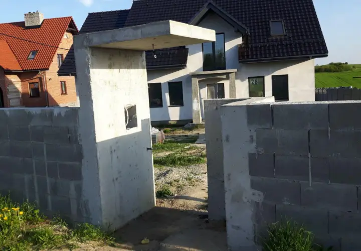 Betonowy daszek nad furtką, budowa ogrodzenia z pustaków, domy w tle.