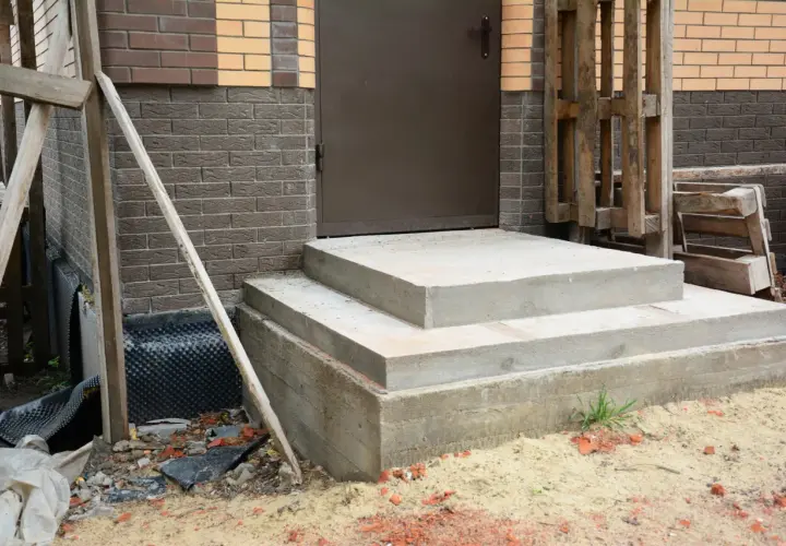 Solidny fundament pod schody z betonu, gotowy na dalsze prace budowlane. Obok widać materiały budowlane i fragment domu.