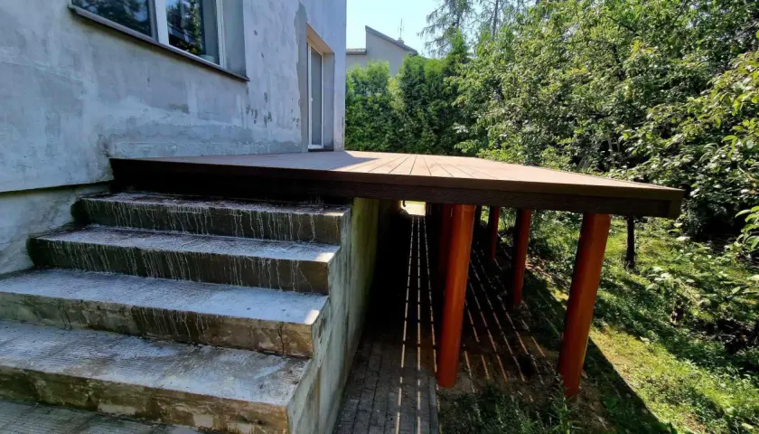 Nowy, wysoki taras na słupach, z drewnianą podłogą, sąsiaduje z betonowymi schodami i zielonym ogrodem.