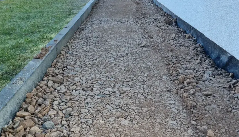 Ścieżka z kruszywa obok trawnika i białej elewacji. Połączenie elewacji z kostką kamienną tworzy estetyczną całość.