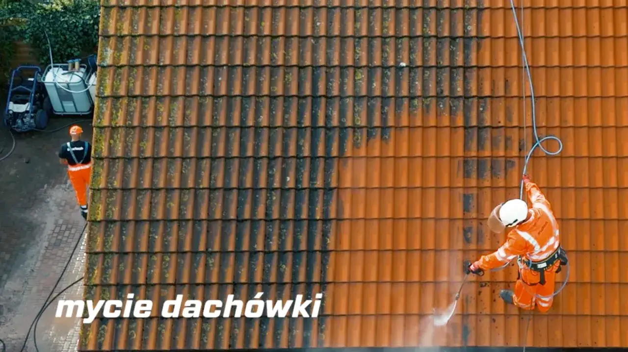 Pracownik w pomarańczowym stroju pokazuje, jak usunąć mech z dachu, czyszcząc dachówkę myjką ciśnieniową.