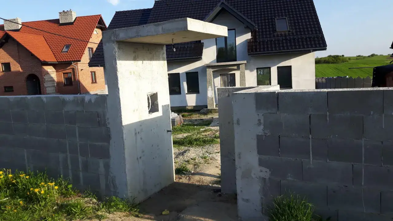 Betonowy daszek nad furtką, budowa ogrodzenia z pustaków, domy w tle.