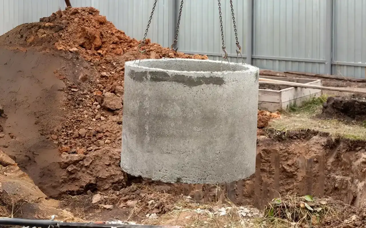 Betonowy krąg unoszony przez łańcuchy nad wykopem. Rozpoczyna się montaż szamba, a cena tej usługi zależy od wielu czynników.