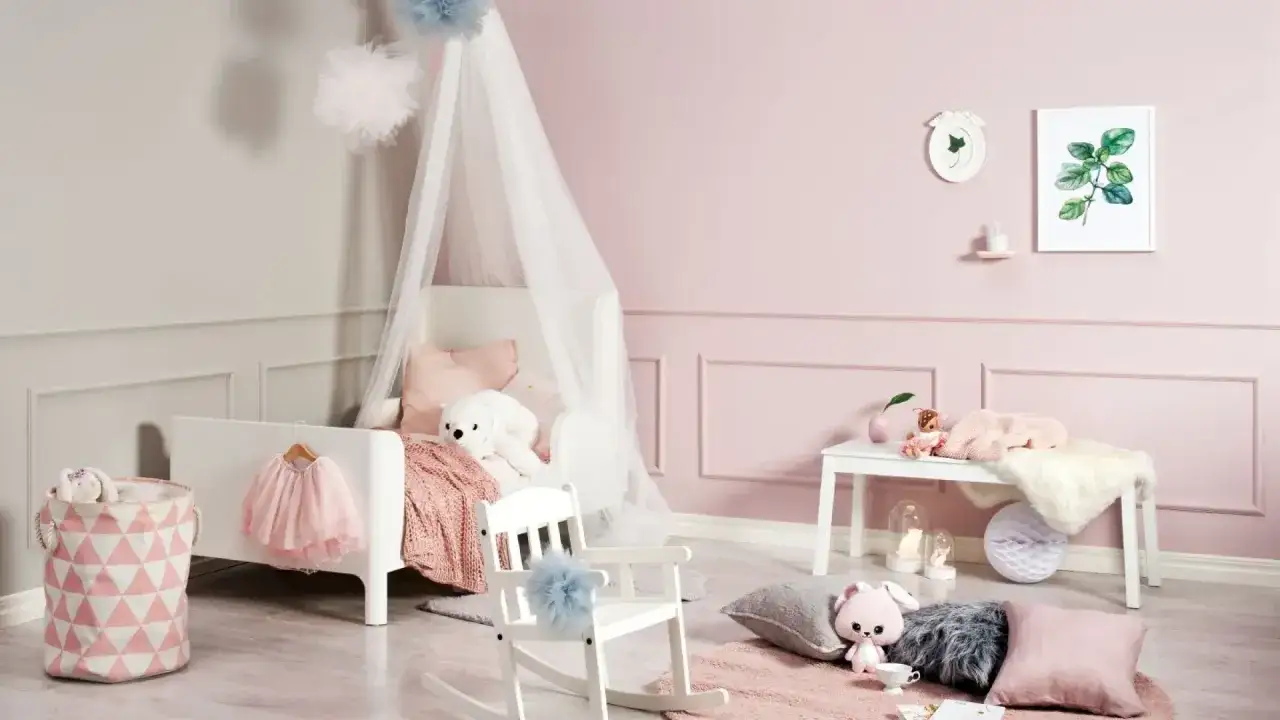 Uroczy pokój dziecięcy w pastelowych kolorach. Widać łóżeczko z baldachimem, huśtawkę, kosz na zabawki i dekoracje. Różnica między Beckers Vaggfarg a designer jest subtelna, ale widoczna w odcieniach różu.
