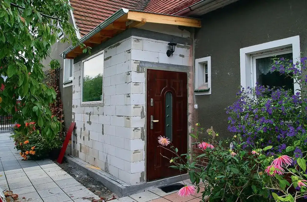 Budowa dobudówki 30m2: białe pustaki, brązowe drzwi, okno, dach, kwiaty.